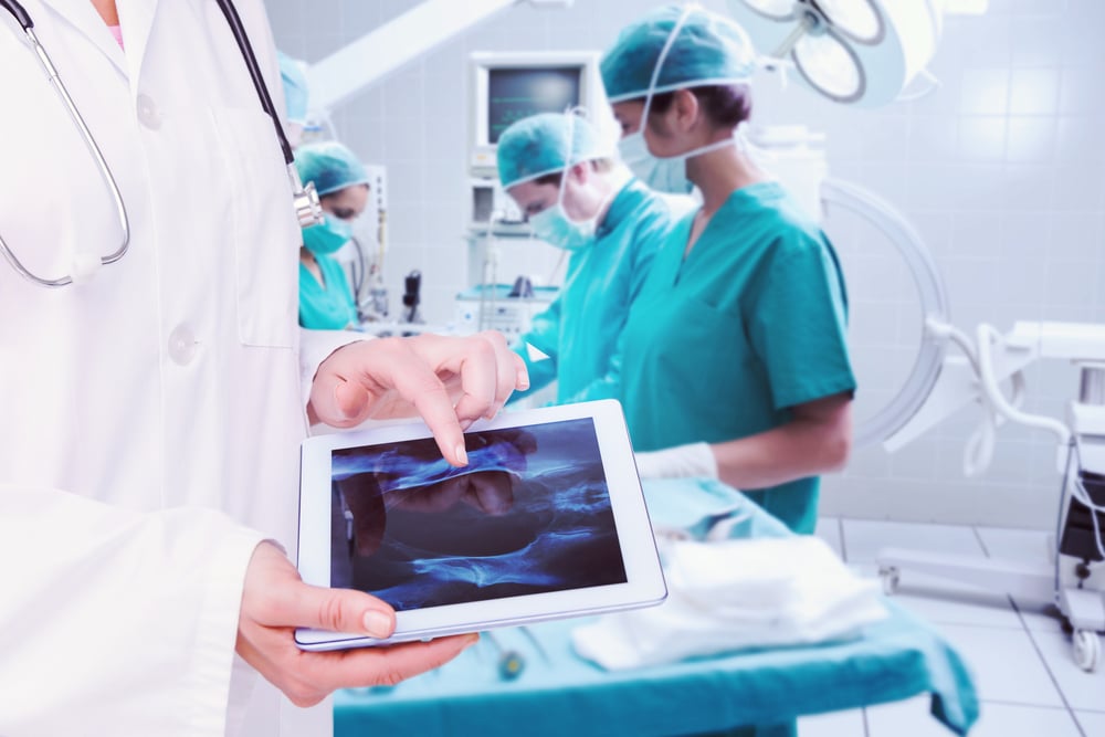 Medico che guarda un'immagine a raggi X su un tablet, sullo sfondo un'équipe che esegue un'operazione.