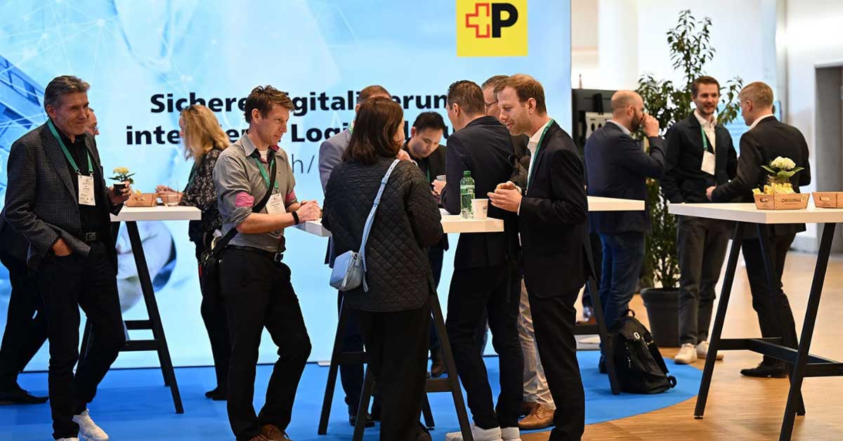 Diverse persone conversano in piedi attorno a tavoli alti in un contesto di fiera o conferenza. Sullo sfondo si vede un grande striscione blu con il logo della Posta e il testo « Soluzione digitale sicura ». Sui tavoli ci sono bevande e piccole decorazioni. Diverse persone conversano in piedi attorno a tavoli alti in un contesto di fiera o conferenza. Sullo sfondo si vede un grande striscione blu con il logo della Posta e il testo « Soluzione digitale sicura ». Sui tavoli ci sono bevande e piccole decorazioni.