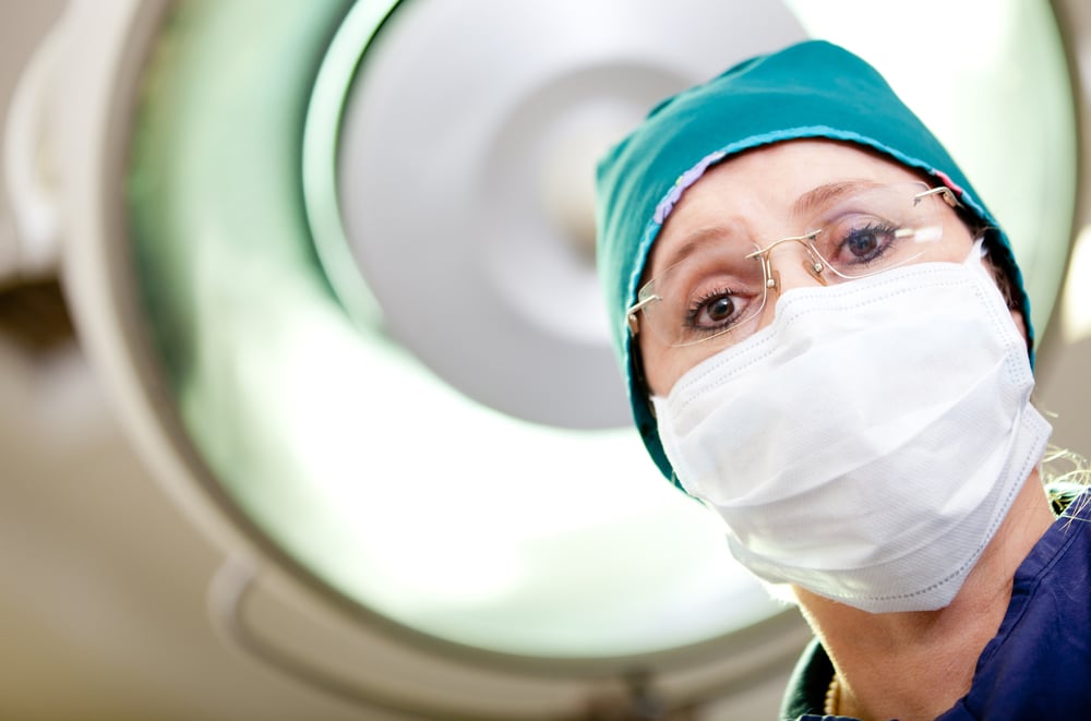 Doctor with surgical mask and glasses looks intently at the patient under the operating light.