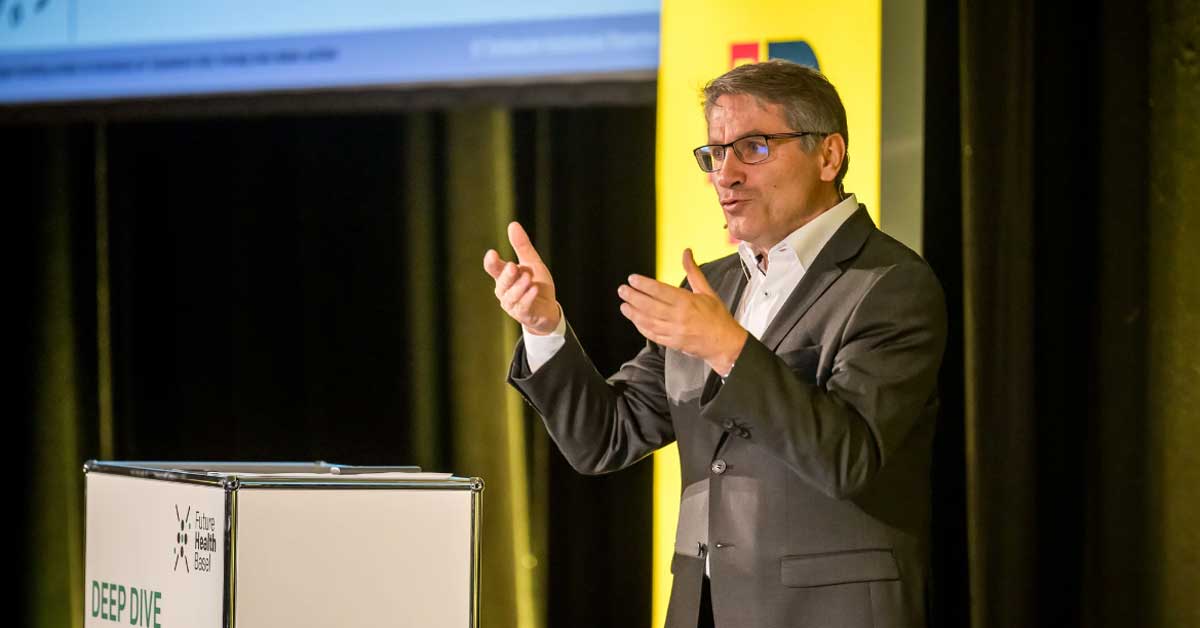 Il Prof. Dr Franz Benstetter parla sul palco e gesticola davanti a un leggio con la scritta « Deep Dive ». Sullo sfondo sono visibili uno striscione giallo e uno schermo. Il Prof. Dr Franz Benstetter parla sul palco e gesticola davanti a un leggio con la scritta « Deep Dive ». Sullo sfondo sono visibili uno striscione giallo e uno schermo.