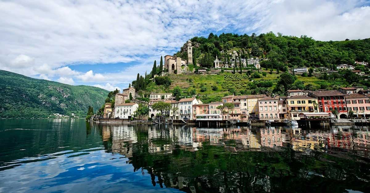 Veduta panoramica di un borgo pittoresco sulle rive di un lago nel Canton Ticino. Diverse case colorate si affacciano sull’acqua, sopra le quali si erge una collina verde con una chiesa e cipressi. Il lago riflette gli edifici e il cielo nuvoloso. Veduta panoramica di un borgo pittoresco sulle rive di un lago nel Canton Ticino. Diverse case colorate si affacciano sull’acqua, sopra le quali si erge una collina verde con una chiesa e cipressi. Il lago riflette gli edifici e il cielo nuvoloso.