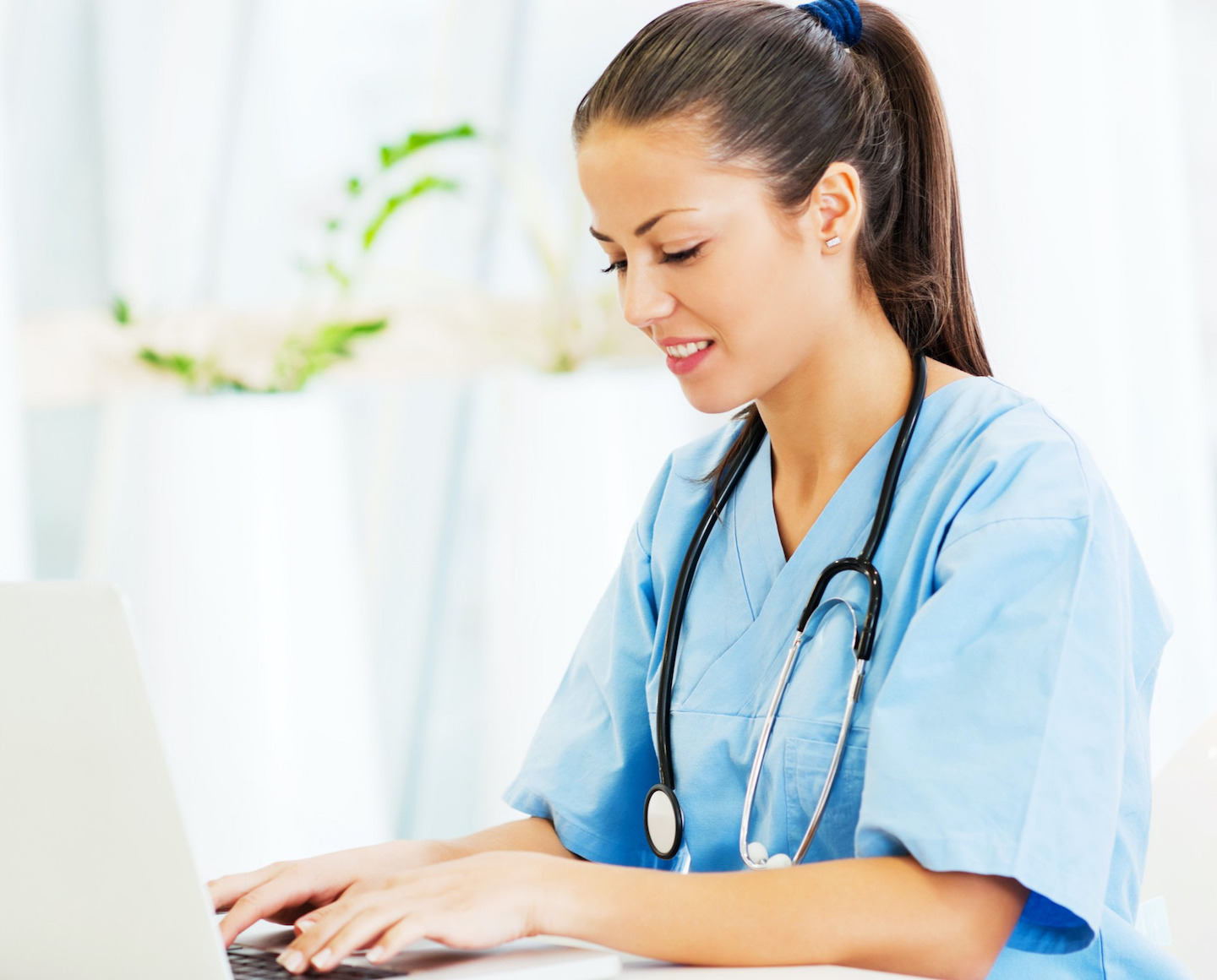 Un'assistente in abiti da lavoro blu con uno stetoscopio al collo lavora su un computer portatile in un ambiente luminoso e accogliente.