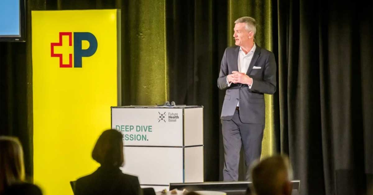 Il Sig. Schneider è sul palco accanto a uno Posta-striscione giallo. Davanti a lui c’è un leggio con la scritta « Deep Dive Session » e il logo « Future Health Basel ». In primo piano sono visibili alcune persone del pubblico. Il Sig. Schneider è sul palco accanto a uno Posta-striscione giallo. Davanti a lui c’è un leggio con la scritta « Deep Dive Session » e il logo « Future Health Basel ». In primo piano sono visibili alcune persone del pubblico.