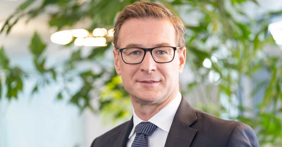 Persona con abito scuro, camicia bianca e cravatta a pois si trova davanti a uno sfondo con piante verdi e luci sfocate. Persona con abito scuro, camicia bianca e cravatta a pois si trova davanti a uno sfondo con piante verdi e luci sfocate.