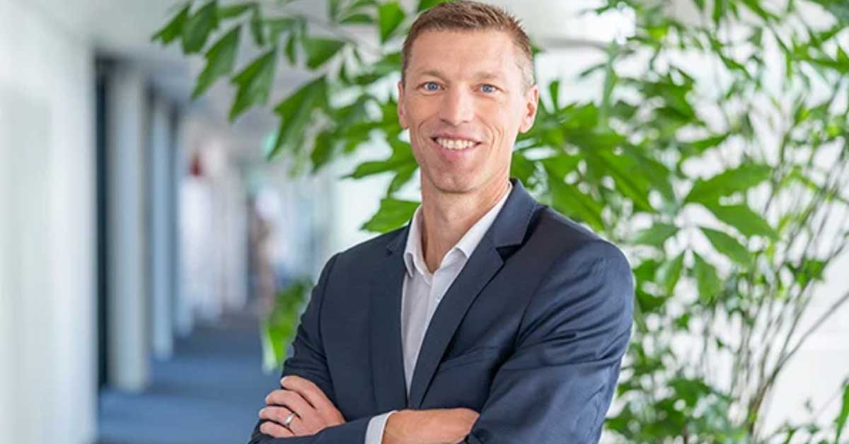 Person wearing a dark blue suit and white shirt stands with arms crossed in a bright, modern hallway. Green plants and blurred architectural details are visible in the background. Person wearing a dark blue suit and white shirt stands with arms crossed in a bright, modern hallway. Green plants and blurred architectural details are visible in the background.